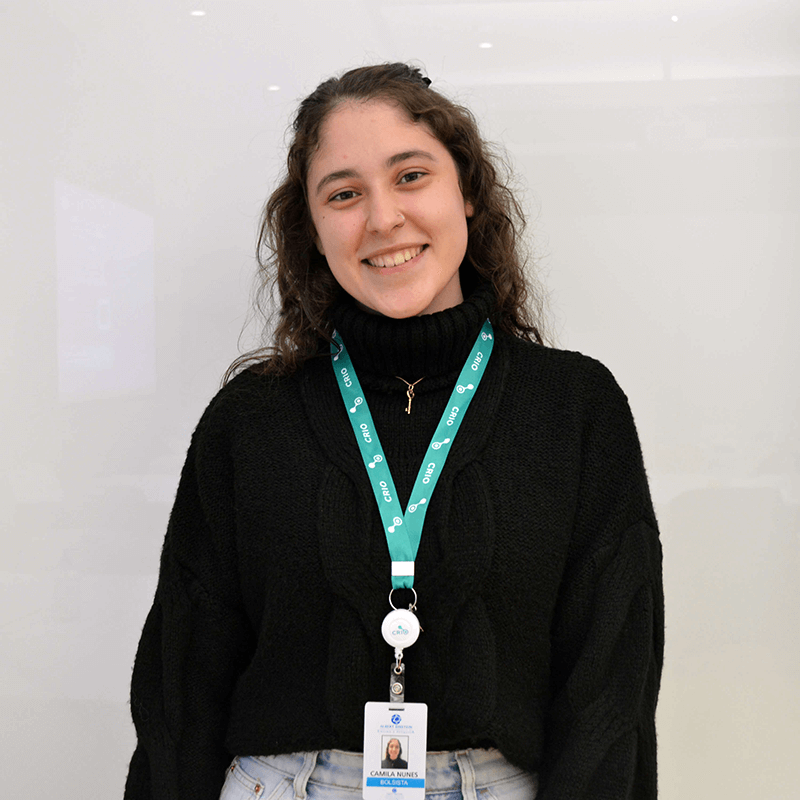 Foto da mestrada do Centro de Pesquisa em Imuno-oncologia, Camila Nunes. Mulher branca, com cabelos castanhos longos, sorrindo para a foto e utilizando blusa preta e crachá do centro de pesquisa.